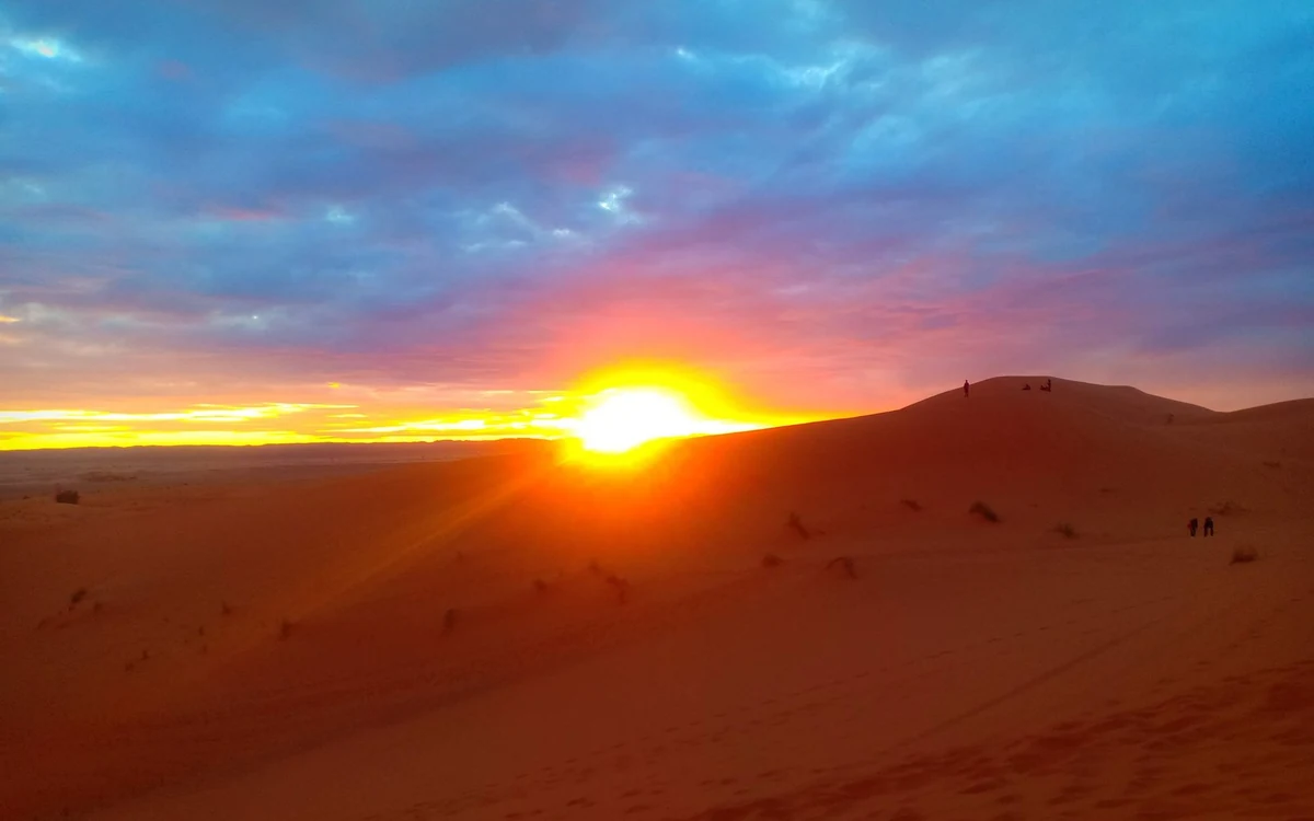 The sun low over a dune ridge, with two figures walking along the crest