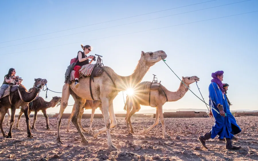 A guide in blue leading camels and riders towards the low sun